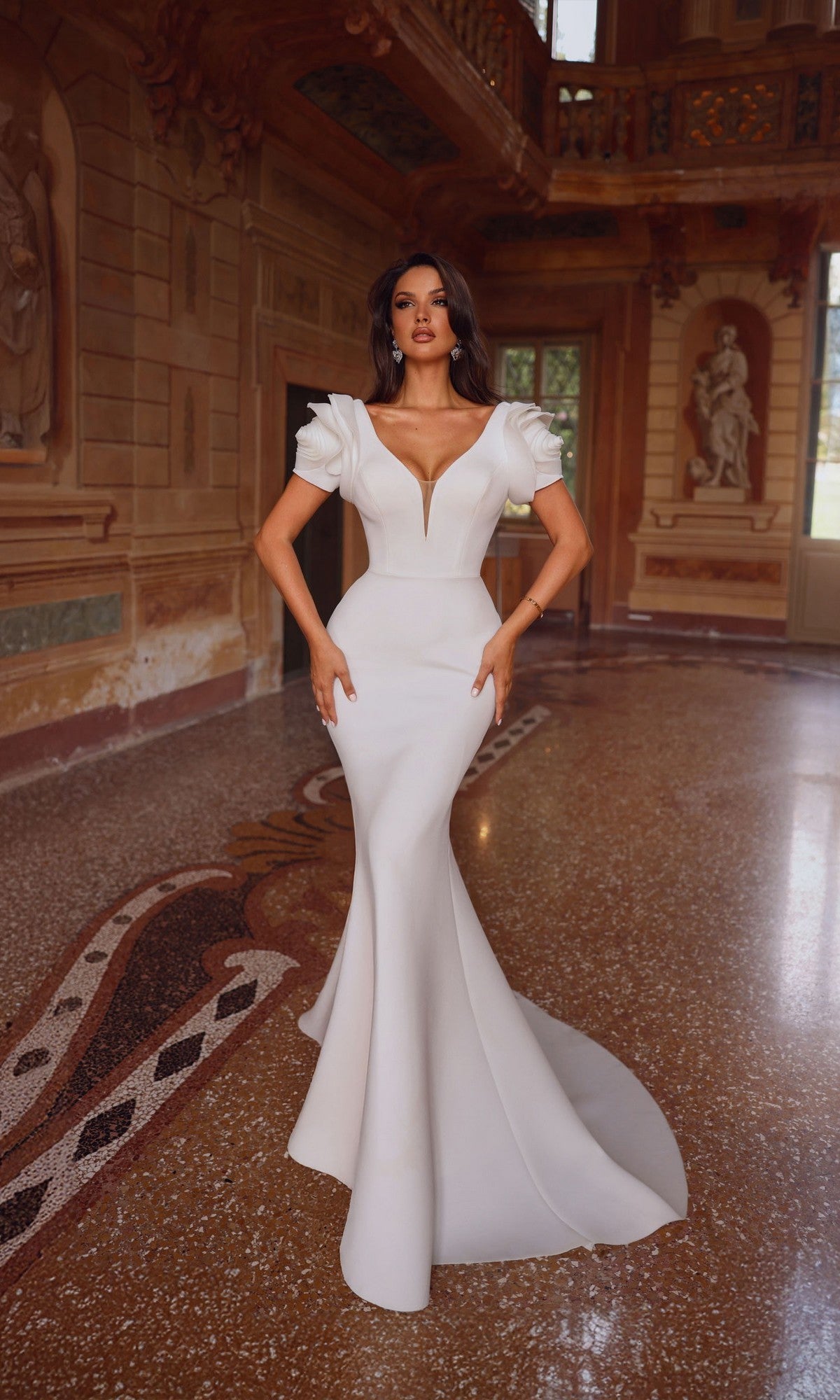 A woman wearing the Alyce 7066 long formal dress in ivory, featuring a fitted silhouette and plunging v-neckline, poses indoors amid ornate classical decor and statues.